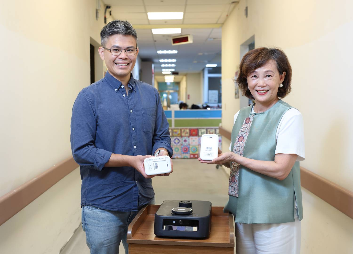 Sigma Robotics founder Snaken (left) and 板橋榮家 Director 陳桂美 (right), holding contactless vital-sign monitors, with Sigma's autonomous mobile robot in front (Image: 經濟部中小及新創企業署 新創採購)